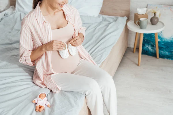 ベッドに座ってベビーシューズを持つ妊婦のトリミングされたビュー — ストック写真
