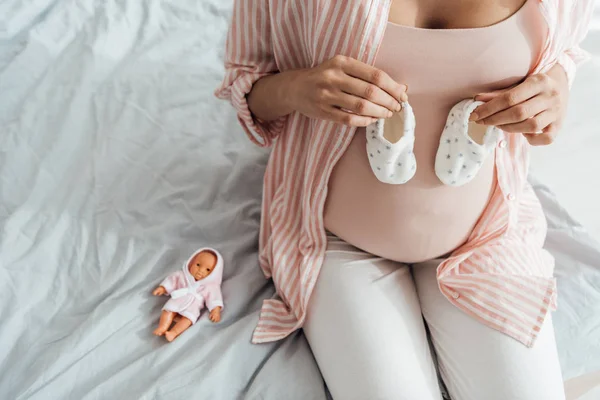 ベッドに座ってベビーシューズを持つ妊婦のトリミングされたビュー — ストック写真