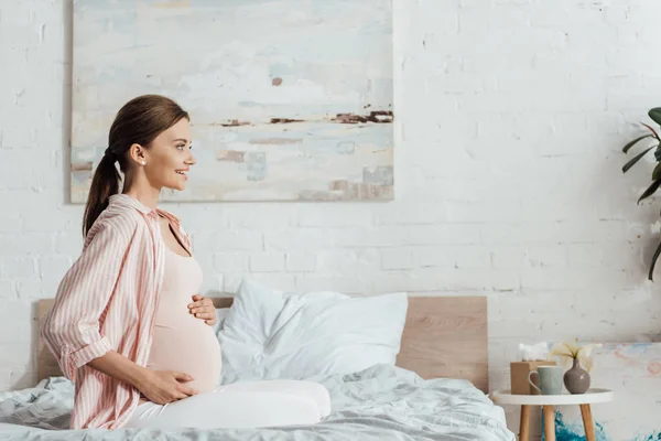Yatakta Oturan Karnına Dokunan Mutlu Hamile Kadının Yan Görünümü — Stok fotoğraf