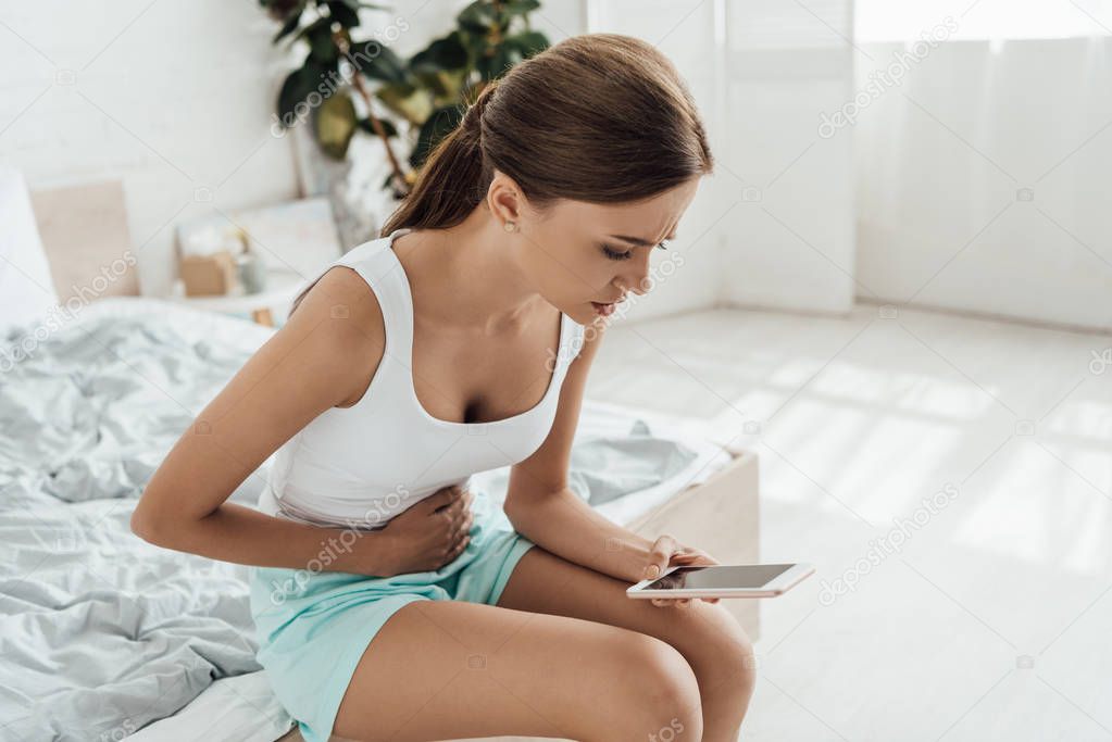 sad young woman touching belly and using smartphone in bedroom