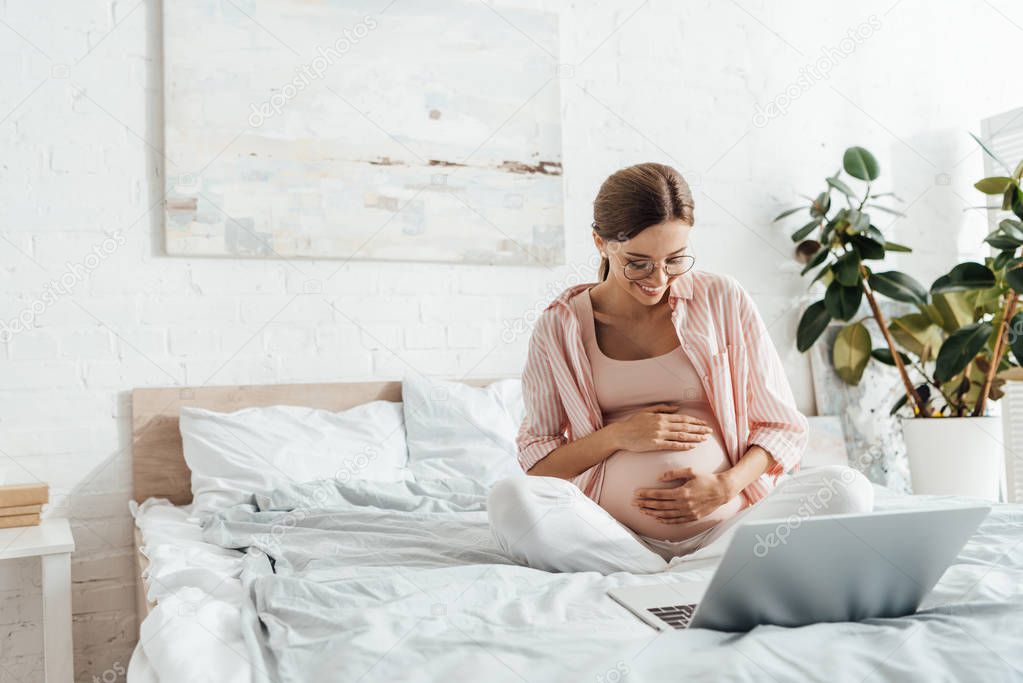 pregnant woman in glasses sitting on bed and using laptop