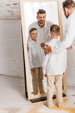 ayna önünde duran beyaz gömlekli gülümseyen baba ve oğulun tam boy görünümü