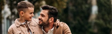 panoramic shot of smiling son and dad embracing and looking at each other in autumn day on street clipart