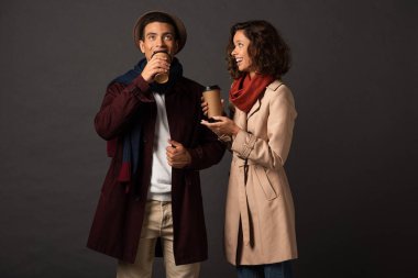 smiling stylish interracial couple in autumn outfit holding coffee to go and talking on black background clipart