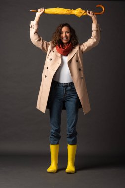 happy woman in trench coat and rubber boots holding yellow umbrella above head on black background clipart