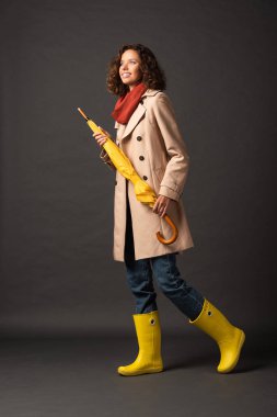 cheerful woman in trench coat and rubber boots holding yellow umbrella on black background clipart