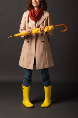 cropped view of smiling woman in trench coat and rubber boots holding yellow umbrella on black background clipart