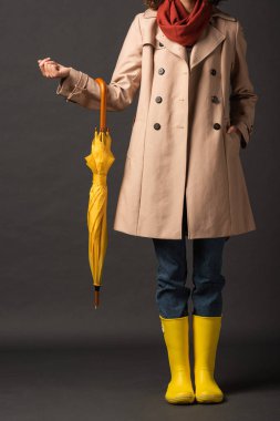 cropped view of woman in trench coat and rubber boots holding yellow umbrella on black background clipart