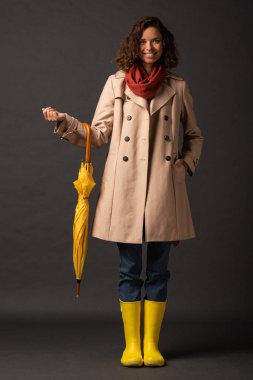 smiling woman in trench coat and rubber boots holding yellow umbrella on black background clipart