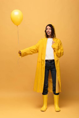 sarı yağmurluk ve wellies sarı arka plan üzerinde balon tutan mutlu kıvırcık kadın