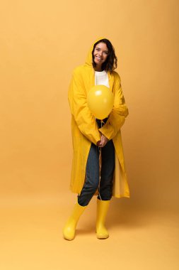 smiling curly woman in yellow raincoat and wellies holding balloon on yellow background clipart