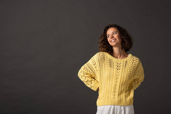 happy curly woman in yellow knitted sweater looking away on black background