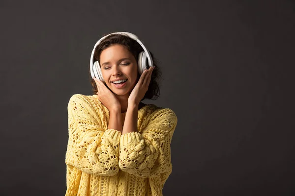 黒い背景にヘッドフォンで音楽を聴く黄色のニットセーターで目を閉じた笑顔の女性 — ストック写真