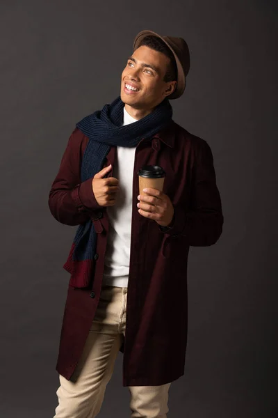 smiling mixed race man in scarf and hat holding paper cup and looking away on black background