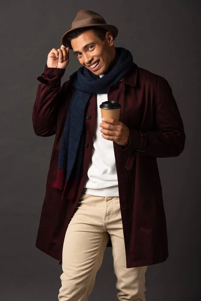 smiling mixed race man in scarf and hat holding paper cup on black background