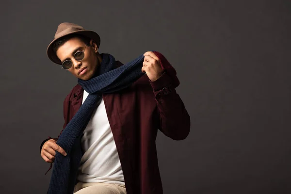 elegant mixed race man in scarf, sunglasses and hat on black background