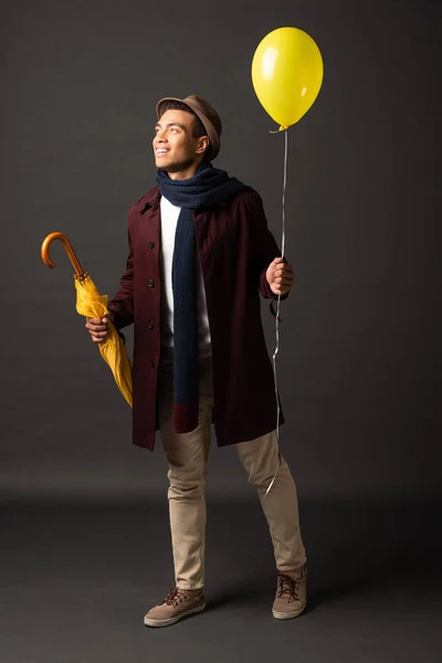 smiling mixed race man in scarf and hat holding yellow balloon and umbrella and looking away on black background