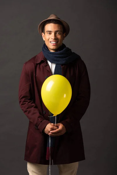 smiling mixed race man in scarf and hat holding yellow balloon on black background