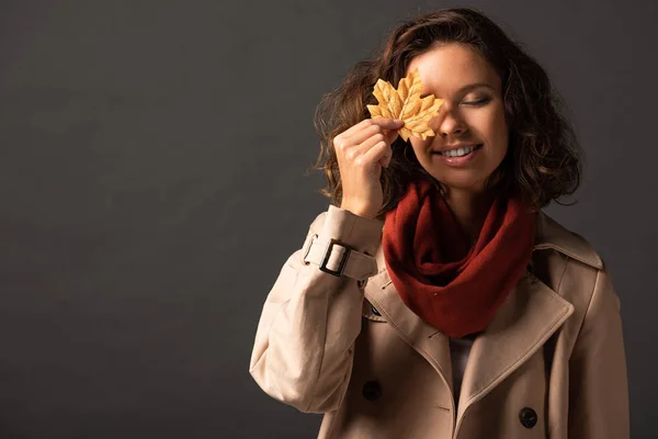 smiling woman in trench coat holding golden maple leaf on eye on black background