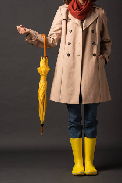 cropped view of woman in trench coat and rubber boots holding yellow umbrella on black background