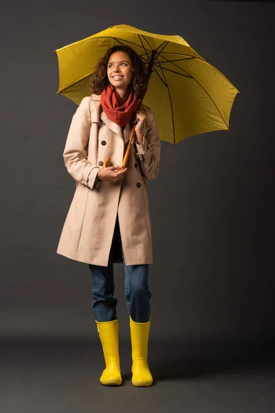 smiling woman in trench coat and rubber boots holding yellow umbrella and looking away on black background