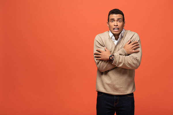 confused mixed race man in beige sweater feeling cold isolated on orange