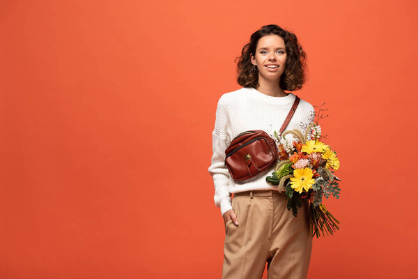 smiling woman in autumnal outfit holding bouquet of flowers isolated on orange
