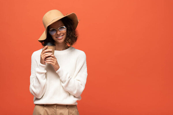 smiling woman in hat and glasses holding coffee to go isolated on orange