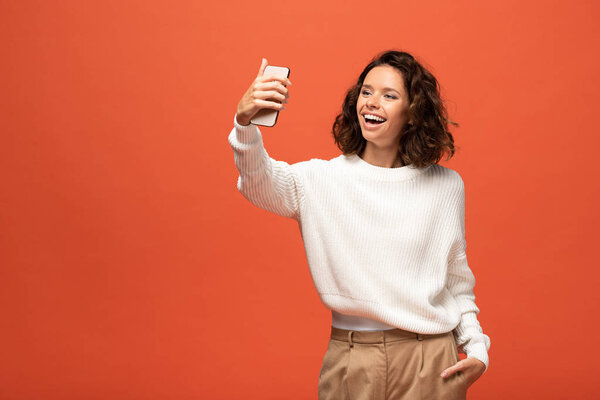 happy woman in autumnal outfit taking selfie with hand in pocket isolated on orange