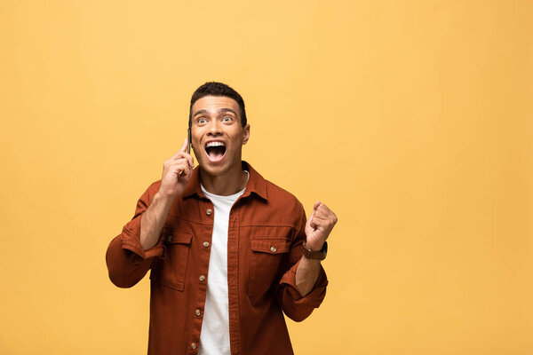 happy mixed race man talking on smartphone and showing yes gesture isolated on yellow