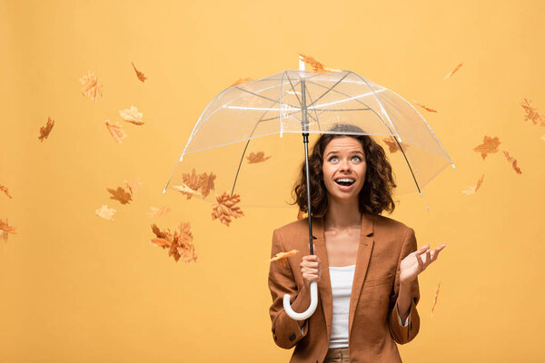 happy curly woman in brown jacket holding umbrella in falling golden maple leaves isolated on yellow