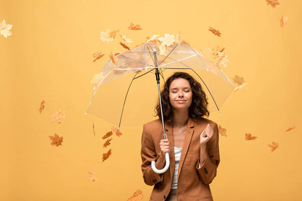 happy curly woman in brown jacket with closed eyes holding umbrella in falling golden maple leaves isolated on yellow