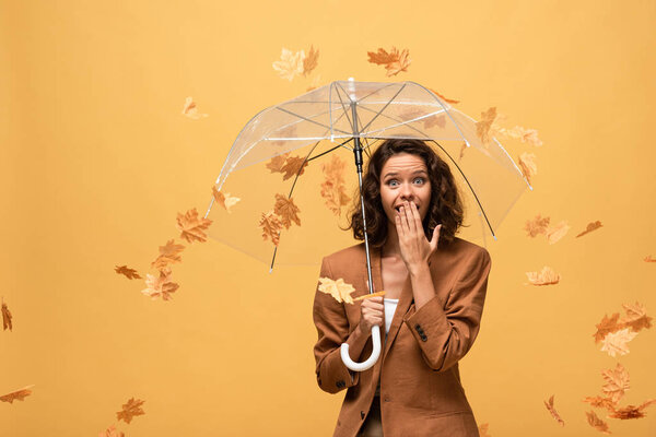 shocked curly woman in brown jacket holding umbrella in falling golden maple leaves isolated on yellow