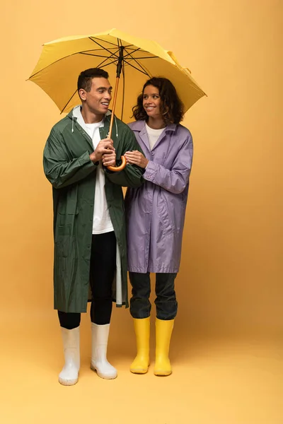 Glimlachend Interracial Paar Regenjassen Rubber Laarzen Houden Paraplu Gele Achtergrond — Stockfoto