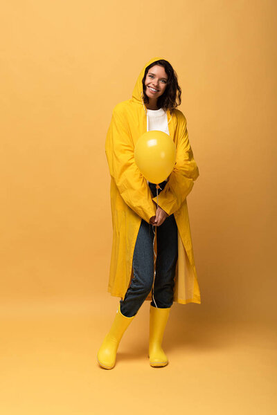 smiling curly woman in yellow raincoat and wellies holding balloon on yellow background