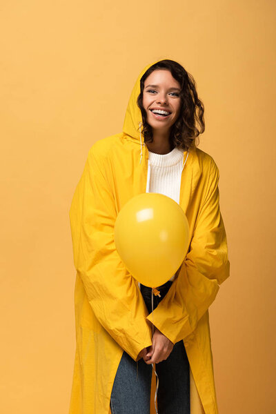 happy curly woman in yellow raincoat holding balloon isolated on yellow