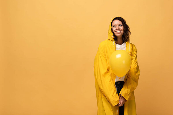 happy curly woman in yellow raincoat holding balloon isolated on yellow