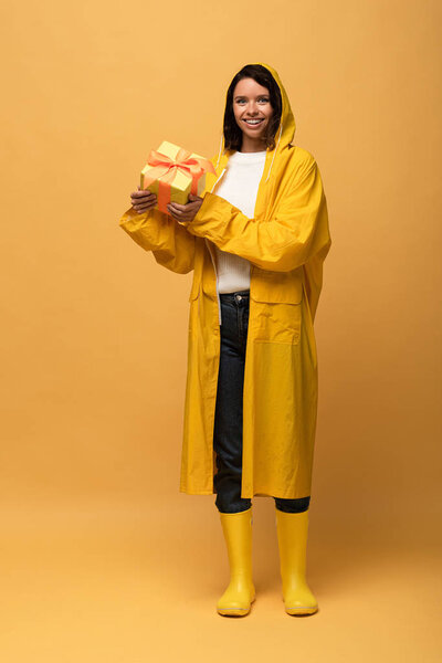 happy woman in yellow raincoat and wellies holding gift box on yellow background