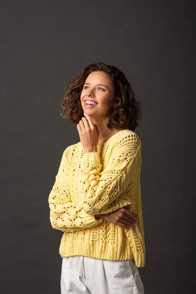 happy curly woman in yellow sweater looking away on black background