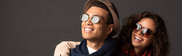 панорамный снимок улыбающейся стильной межрасовой пары в осеннем наряде, смотрящей в сторону на черном фоне
