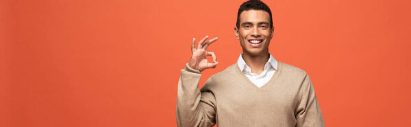 Панорамный снимок счастливого мужчины смешанной расы в бежевом свитере, показывающий знак ОК, изолированный на оранжевом
