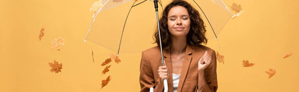 panoramic shot of happy curly woman in brown jacket with closed eyes holding umbrella in falling golden maple leaves isolated on yellow