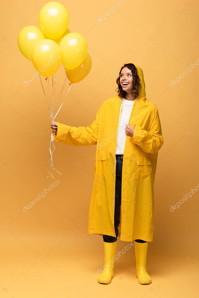 Happy curly woman in yellow raincoat and wellies holding balloons on yellow background