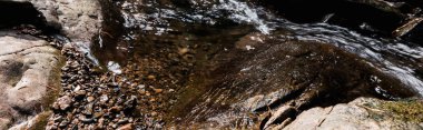 panoramic shot of sunshine on flowing river near wet stones in park  clipart