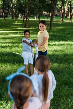 selective focus of cheerful multicultural kids competing in tug of war outside  clipart
