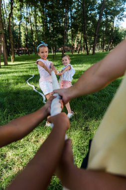 cropped view of multicultural children competing in tug of war with cute friends outside  clipart
