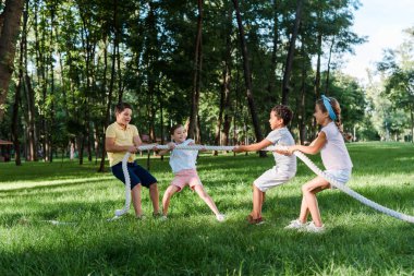 cheerful multicultural kids competing in tug of war outside  clipart