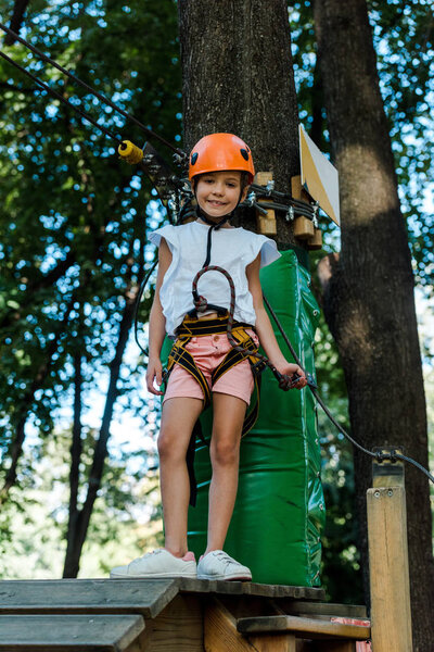 cheerful kid in helmet with height equipment in adventure park near trees 