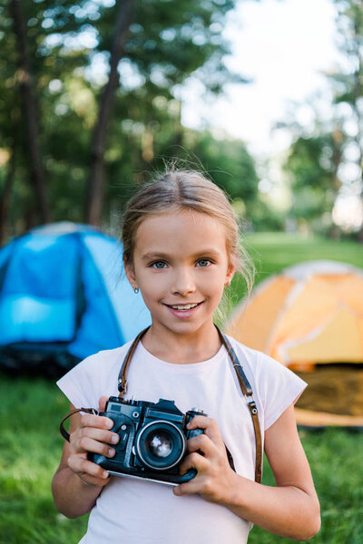 happy kid holding digital camera and smiling near camps 
