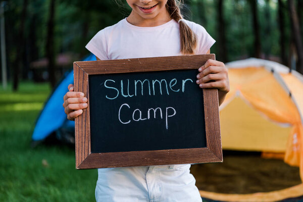 cropped view of happy kid holding chalk board with summer camp letters 